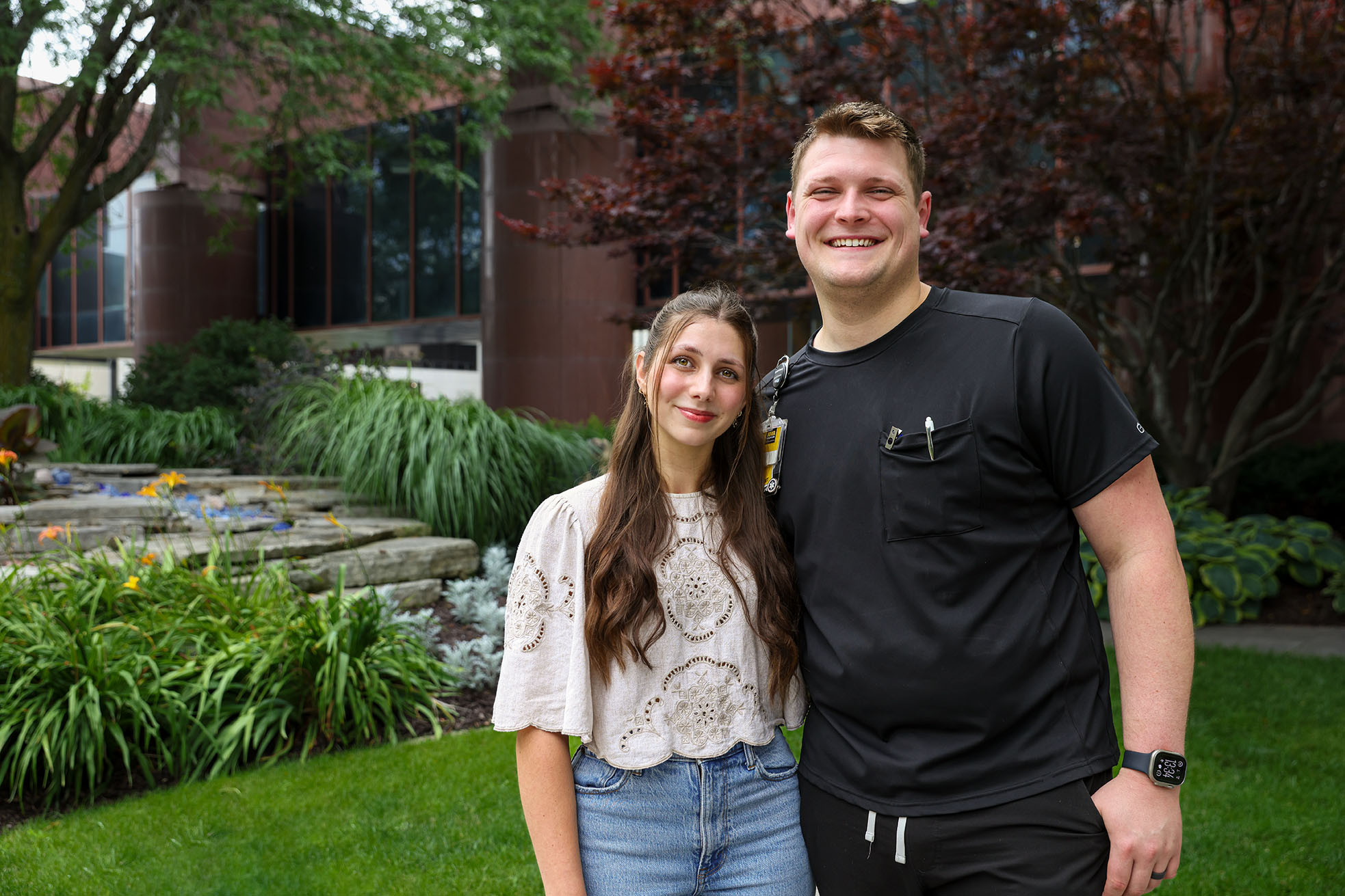 Woman in white shirt and man in black scrubs smile for a photo outside a building. 