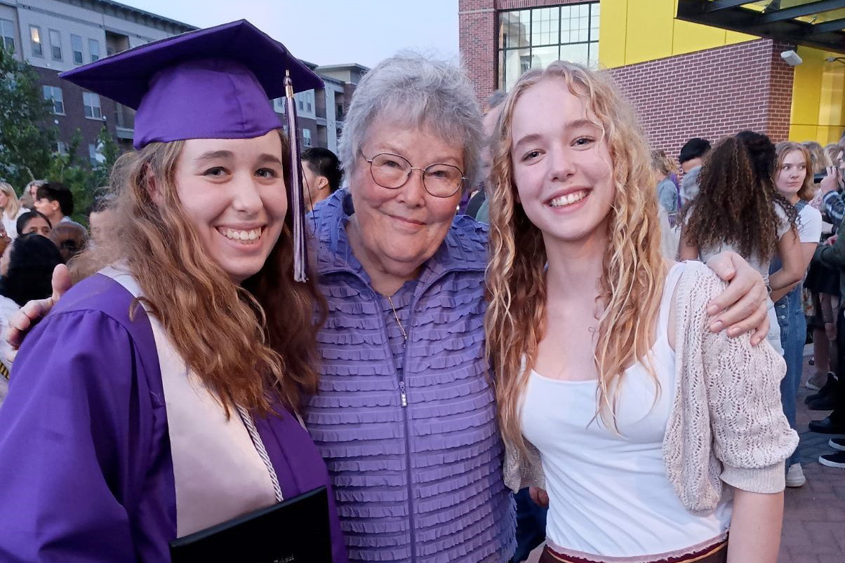 Older woman in purple stands in middle with arms around two smiling younger women, one wearing a purple graduation cap and gown. 