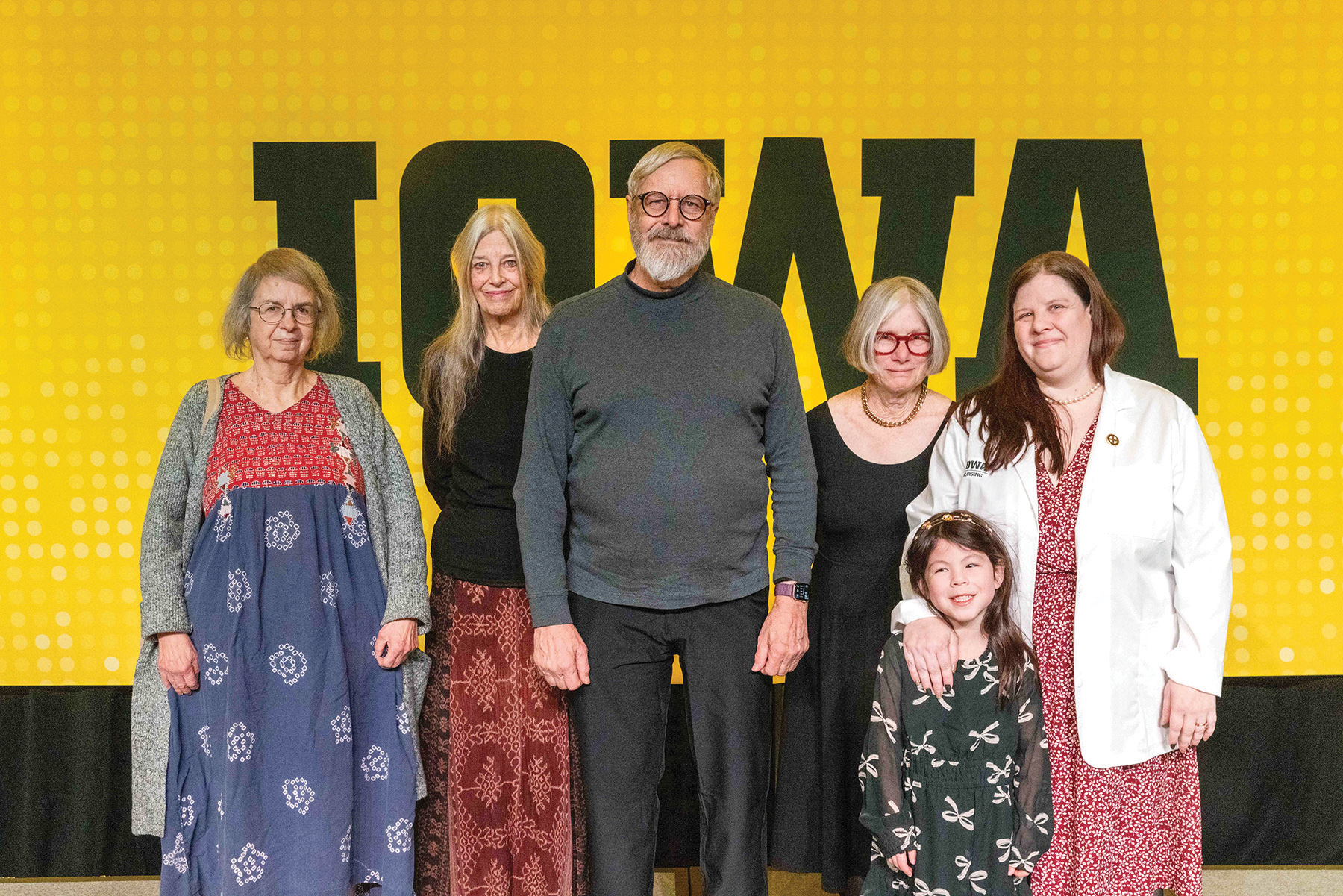 Four adults of varying ages, one wearing a white coat, and a child stand in front of a black and gold Iowa logo banner.