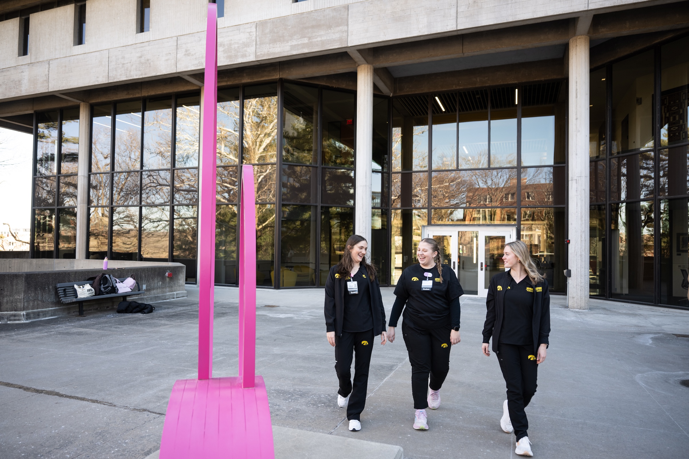 Three people in black scrubs chat as they walk towards the camera. Behind them is a building with large windows on the first floor. To their right is a pink sculpture. 