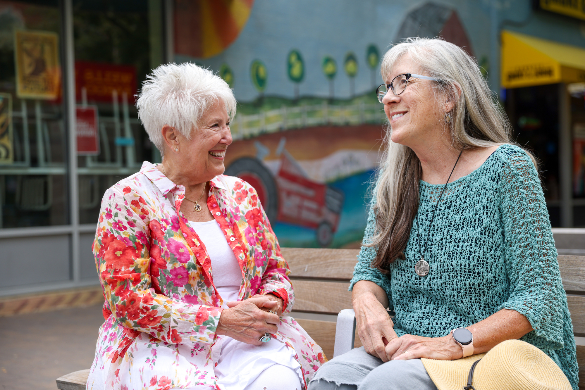 Two older people sit on bench chatting.