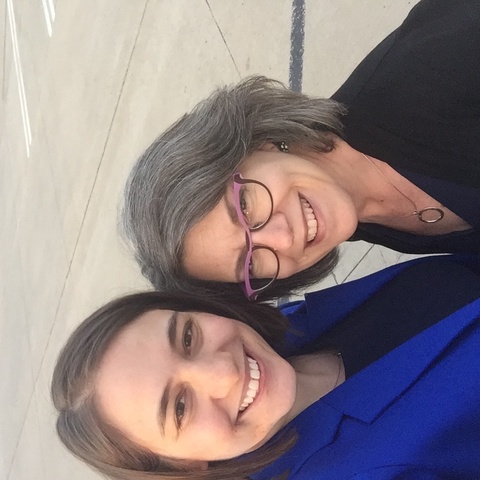 Woman with long hair and blue blazer stands next to shorter woman in black. Photo is a selfie, taken from above. both are smiling. 