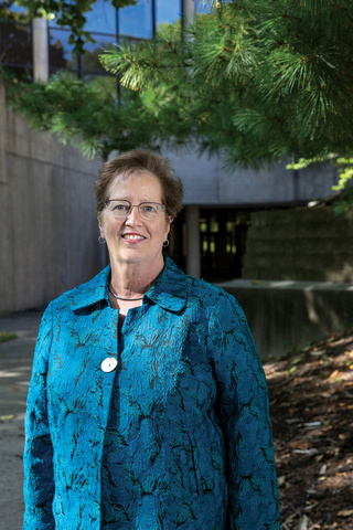 Julie Zerwic portrait. Woman in blue jacket stands outside building and trees smiling