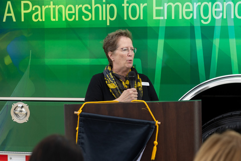 Woman in a black shirt and black and gold scarf holding a microphone stands at a podium talking.