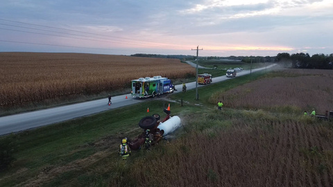 In the foreground is a tractor and white cylindrical trailer lying on their side. Two emergency personnel stand next to the tractor. A road runs through the middle, at a diagonal. On the road are two fire trucks and a SIM-IA truck. Cornfields and sky fill the background. 