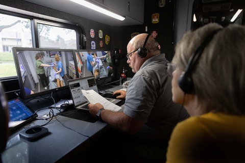 A man wearing headphones looks down at a paper he holds, his hand on a mouse. In front of him is a laptop and two large monitors. On the monitors are people in scrubs working with a figure in a hospital gown lying on a gurney. 