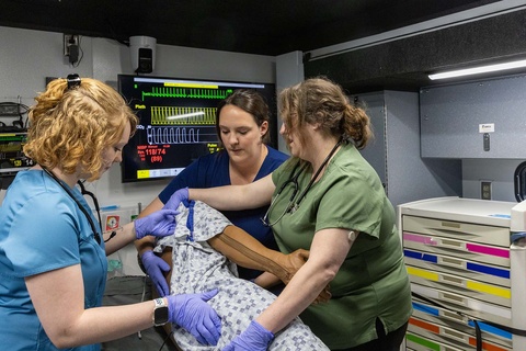 In a small area with medical equipment and a screen on the wall showing readings, three people in scrubs turn a manikin wearing a hospital gown on its side.  