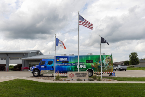 A large blue and green truck is parked behind a sign for the Alburnett Otter Creek Fire Department and three flying flags. Behind is is a low gray building. Above is a large expanse of clouds in the sky. 