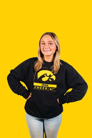 A woman wearing a black Iowa Cheer sweatshirt stands with both hands on hips,, smiling at camera. The background behind her is yellow.