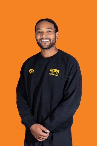 A man in a black sweatshirt with hands crossed casually in front of him poses smiling at the camera. Background behind him is orange.