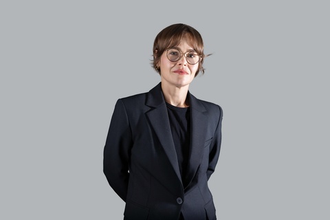 A woman in a black blazer and hat poses with her arms behind her back. The background behind her is gray.