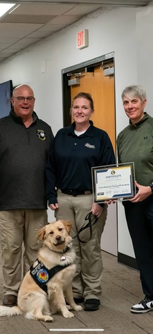 Three people stand together, one holding a leash with a dog sitting in front of her. Person on the right holds a certificate.