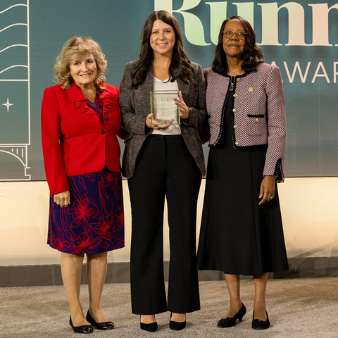 Three people standing on stage at an award ceremony, with the center person holding a glass award.