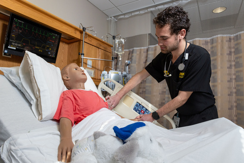 A man in black scrubs with tends to a simulated patient manikin in a hospital room setting. 