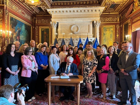 Man sits at desk writing surrounded by a large group of people. 