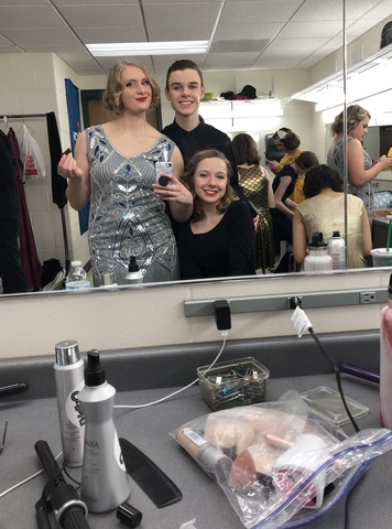 One person in a silver dress and two people in black pose for a selfie in a large mirror. Behind them people are getting ready, and in front of them on the counter are makeup and hair supplies