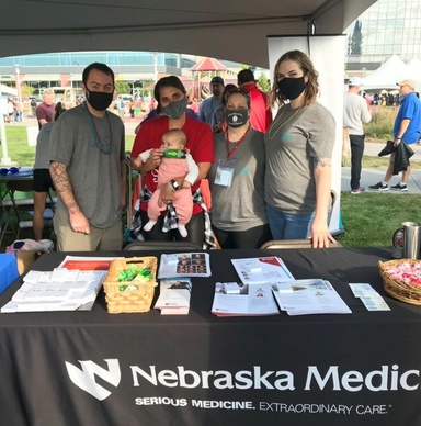 Four people wearing masks and a baby pose for a photo behind a table covered in handouts. 