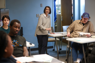 Woman standing at classroom desk looking up, two people in front of her at desks looking at papers