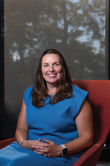 Woman in blue dress sits in orange chair smiling at camera.