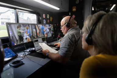 Two screens show people in scrubs working with a simulation manikin. Viewer sees the back of a man with a laptop in front of the screens