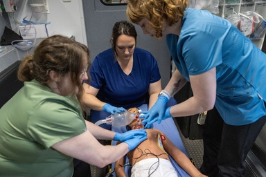 Three people in scrubs surround a simulation manikin, two sitting and on standing. One holds a bag attached to a tube over the patient's mouth while the others appear to be doing chest compressions.