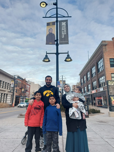 A woman holding a baby stands next to a man who has his hands on the shoulders of two boys standing in front of him. Behind the group is a pole with a banner at the top. On the banner is a photo of a woman.