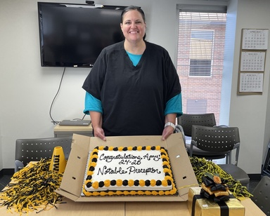 A woman in a black scrub top with a blue shirt underneath smiles as she holds up a cake with black white, and gold frosting that reads "congratulations Amy! 24-25 Notable Preceptor.