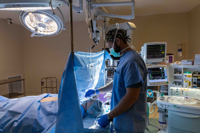 A man in blue scrubs stands in an operating room. A patient on a bed is in front of him. with a blue sheet rising from their neck area. His hand is on an mask that covers the patient's nose and mouth.