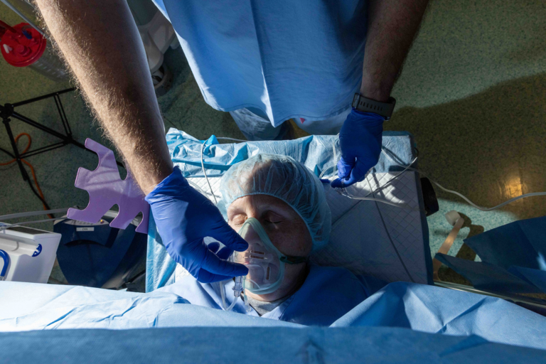 Photograph taken from above. A person with eyes closed wearing a surgical cap lies on a bed in an operating room. A gloved hand is adjusting the clear plastic mask that covers the patient's nose and mouth.