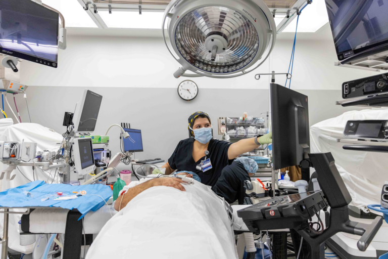 A person in black scrubs with a hap and gown on, and a tag that reads Anesthesia, reaches to adjust something on a computer screen. In front of her is a patient who appears to be under sedation, lying on a bed covered with a white sheet.
