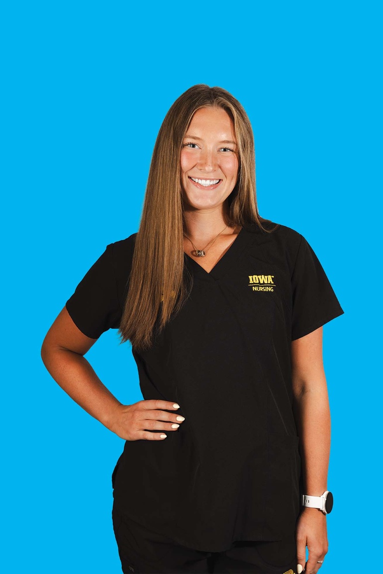 A woman wearing black scrubs poses with one hand on hip, smiling at camera. The background behind her is blue..
