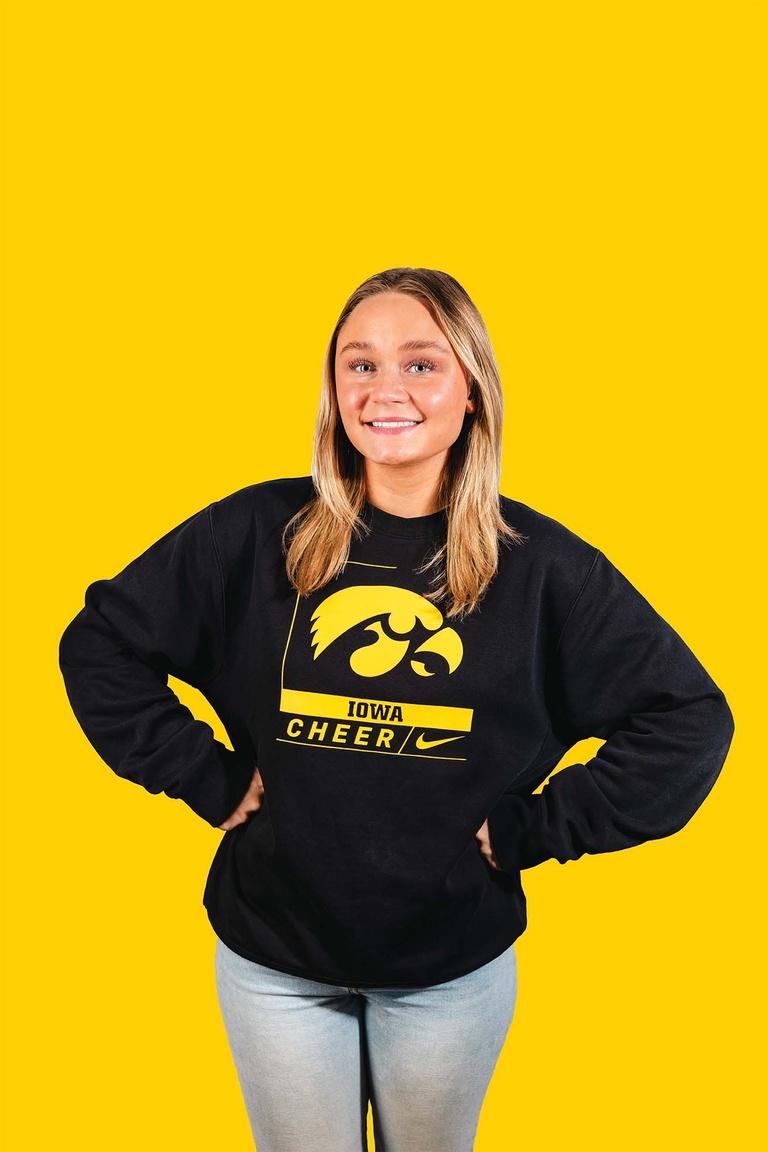 A woman wearing a black Iowa Cheer sweatshirt stands with both hands on hips,, smiling at camera. The background behind her is yellow.