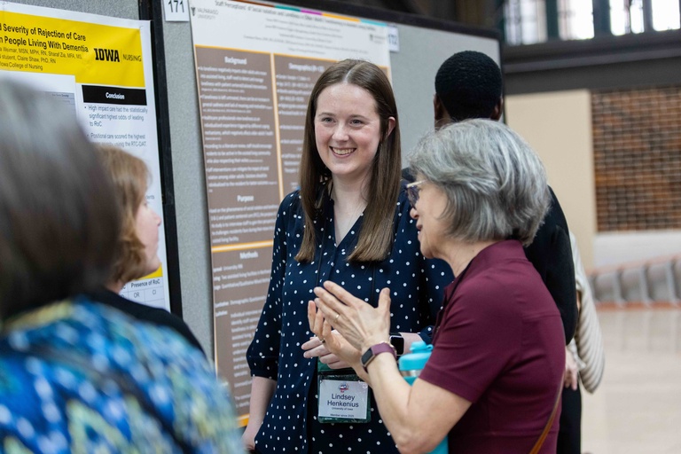 Smiling woman stands in front of scientific poster. Three people are around her talking, not facing camera. 