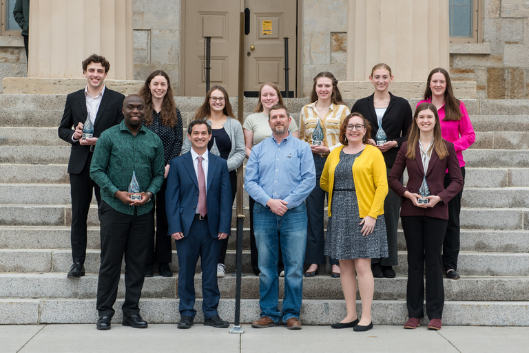 Group of smiling people, most holding glass statues, stand in front of collegiate building., 
