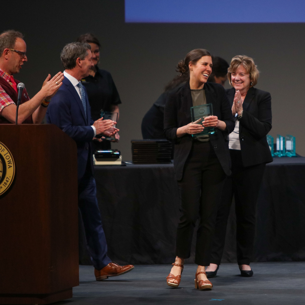 Smiling woman on stage with award, surrounded by clapping and smiling people.