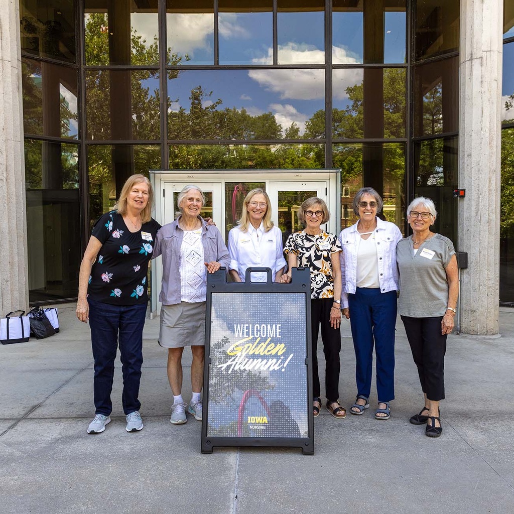 Group of five smiling people stand around a sign that reads 'welcome golden alumni'. Behind them is the entrance to a building.