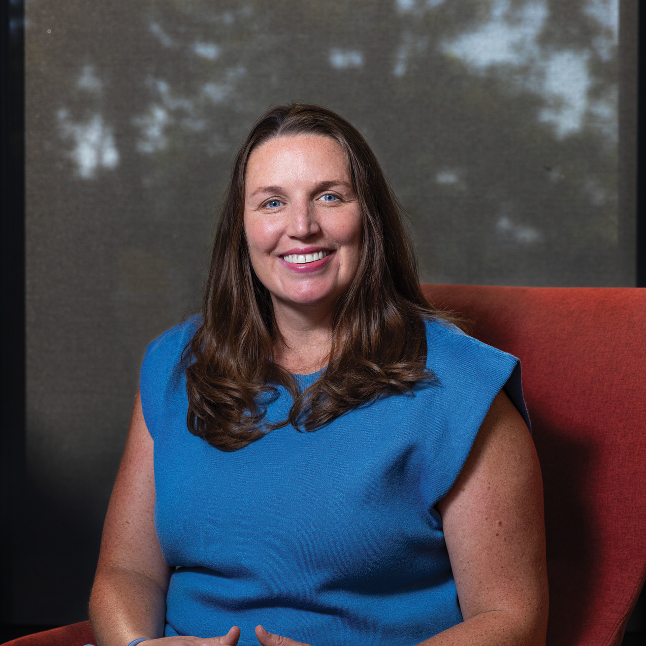Woman in blue dress sits in orange chair smiling at camera.