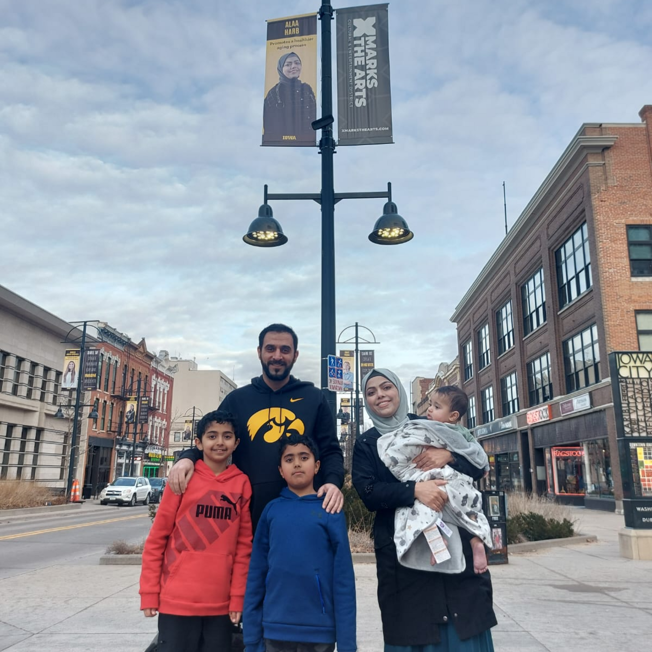 A woman holding a baby stands next to a man who has his hands on the shoulders of two boys standing in front of him. Behind the group is a pole with a banner at the top. On the banner is a photo of a woman.