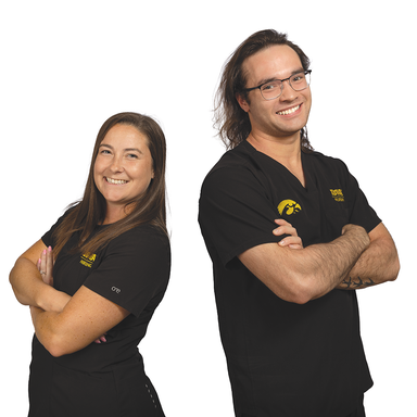 Two students in black scrubs stand with their backs to each other, arms crossed and smiling. The background is white.