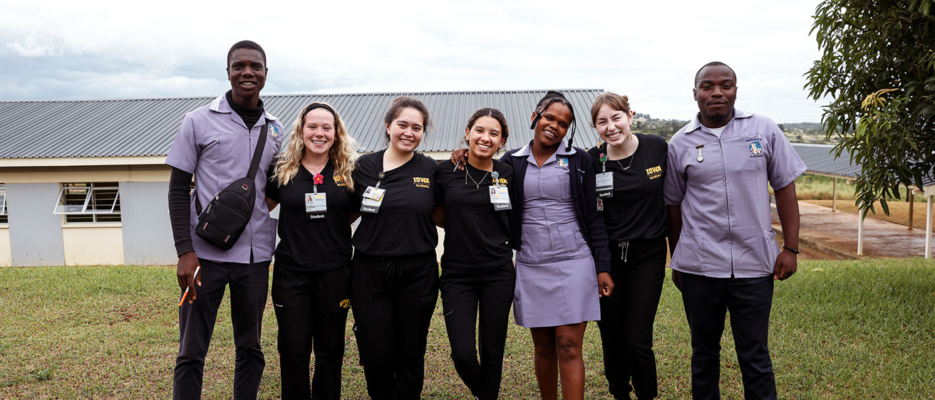 Group of six people with arms around shoulders smile at camera. They are standing outside. Four are in Iowa Nursing scrubs.