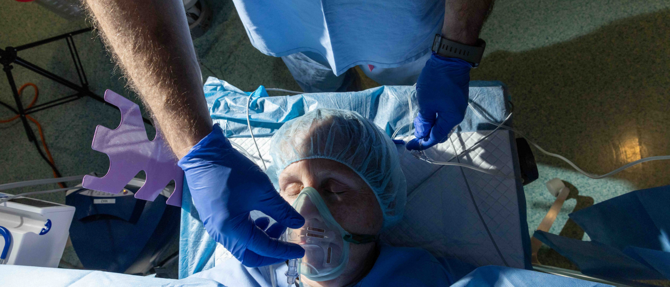 Photograph taken from above. A person with eyes closed wearing a surgical cap lies on a bed in an operating room. A gloved hand is adjusting the clear plastic mask that covers the patient's nose and mouth. 