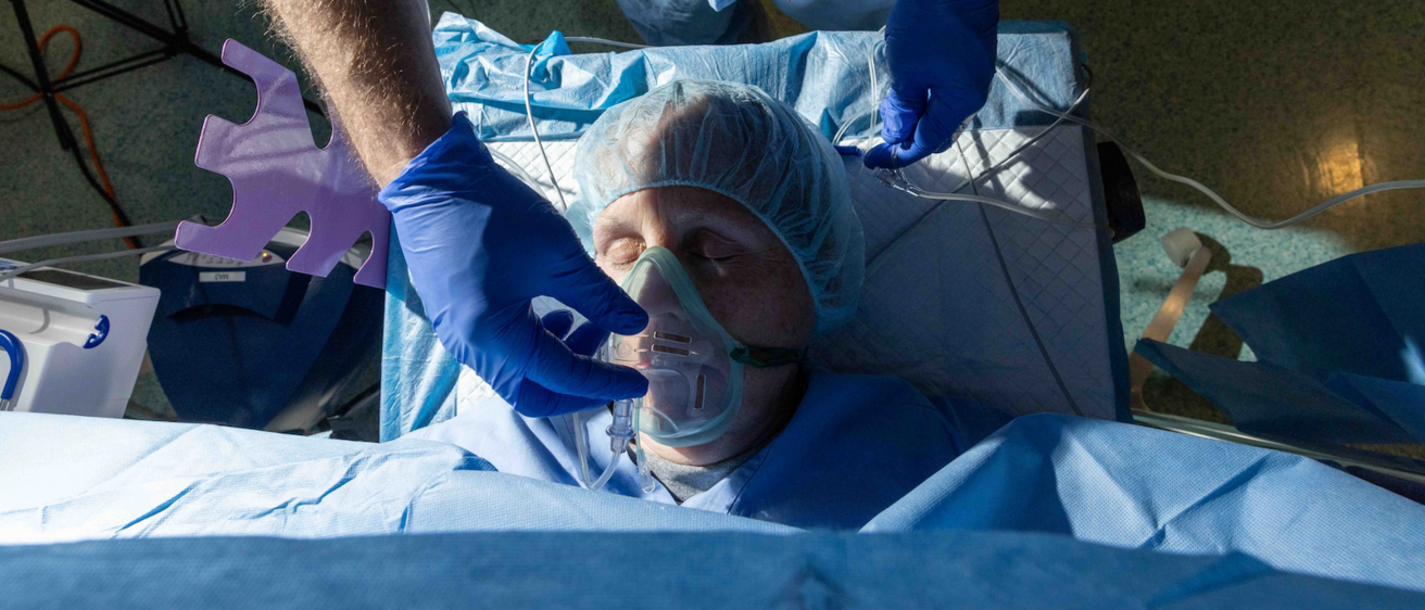 Photograph taken from above. A person with eyes closed wearing a surgical cap lies on a bed in an operating room. A gloved hand is adjusting the clear plastic mask that covers the patient's nose and mouth. 