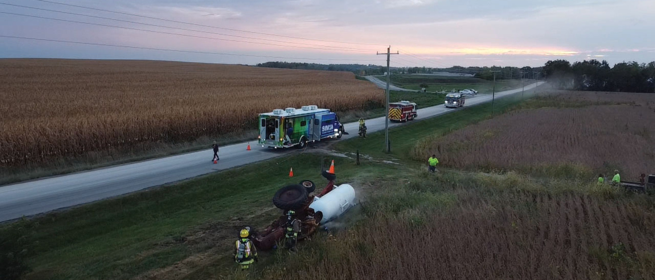 In the foreground is a tractor and white cylindrical trailer lying on their side. Two emergency personnel stand next to the tractor. A road runs through the middle, at a diagonal. On the road are two fire trucks and a SIM-IA truck. Cornfields and sky fill the background. 