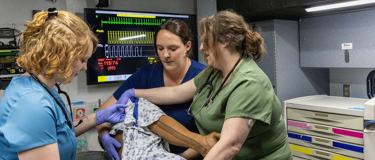 In a small area with medical equipment and a screen on the wall showing readings, three people in scrubs turn a manikin wearing a hospital gown on its side.  