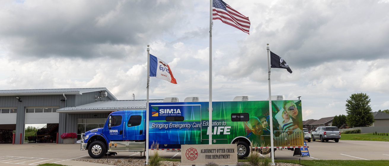 A large blue and green truck is parked behind a sign for the Alburnett Otter Creek Fire Department and three flying flags. Behind is is a low gray building. Above is a large expanse of clouds in the sky. 