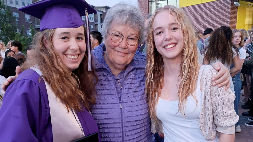 Older woman in purple stands in middle with arms around two smiling younger women, one wearing a purple graduation cap and gown. 