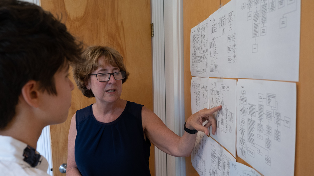 Barb St. Marie stands in front of a board full of papers, pointing and something and talking to a person whose back we see. 