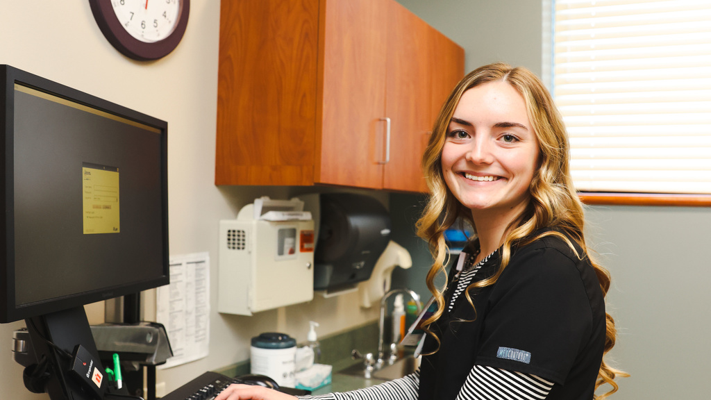 woman in scrubs stands at computer and smiles at camera.