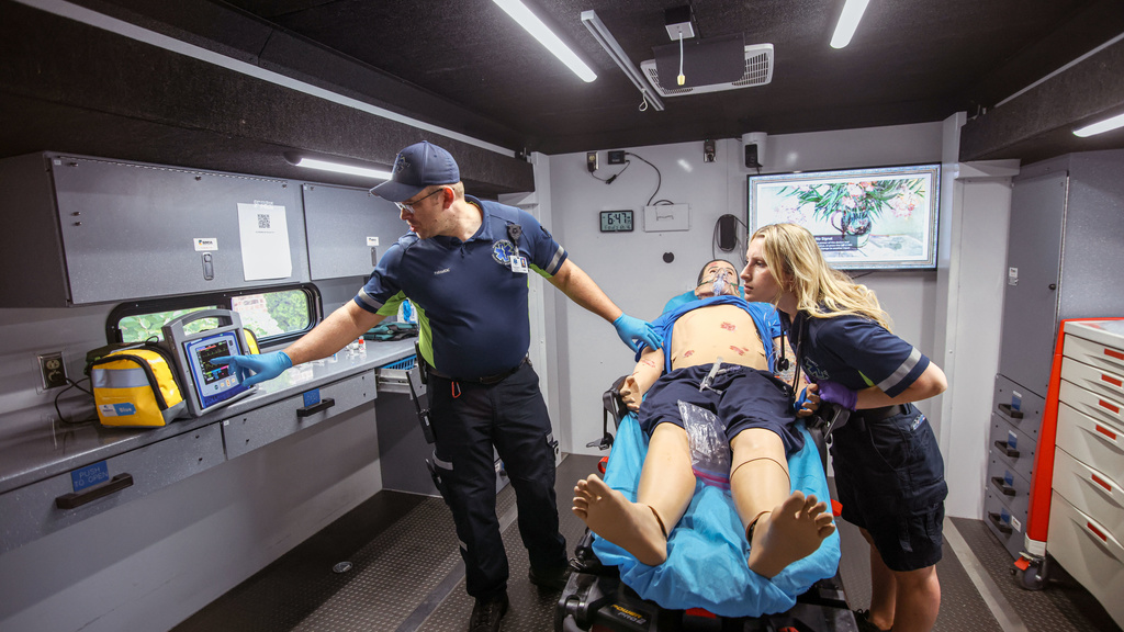 Two paramedics look at machine while working with a manikin in a space that mimics an ambulance bay.