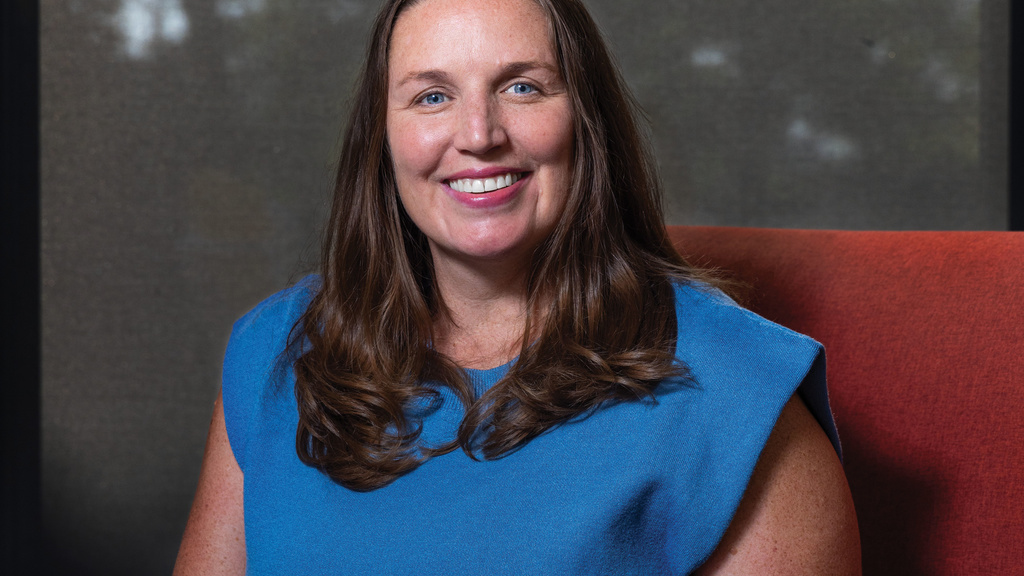 Woman in blue dress sits in orange chair smiling at camera. 